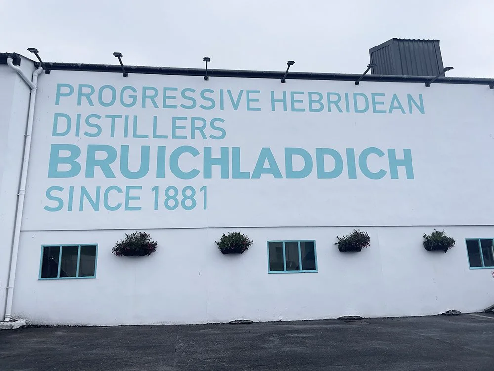 Large exterior wall displaying text about Progressive Hebridean Distillers, known as Bruichladdich since 1881, with three flowering planters and small windows below.