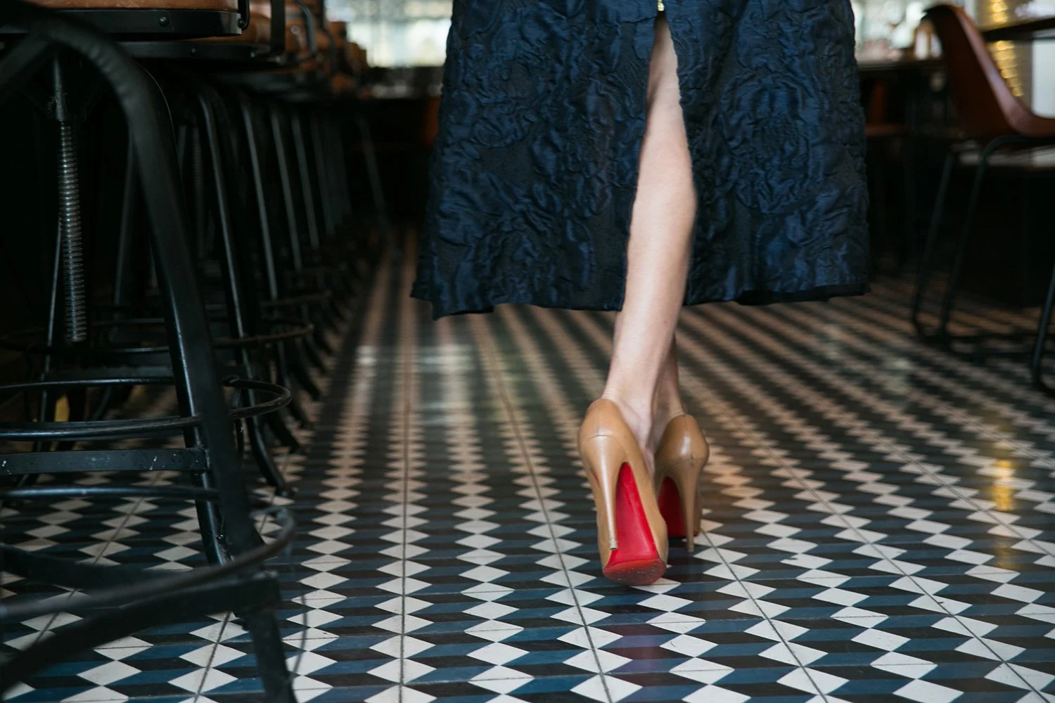 A person walking in a restaurant or cafe wearing high-heeled shoes and a dark blue skirt with a slit, revealing their leg.