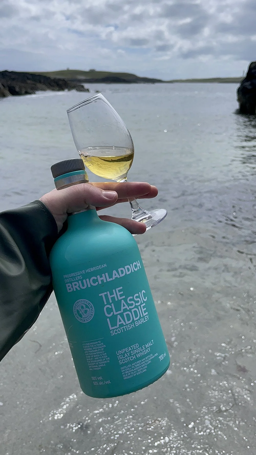 A hand holding a bottle of Bruichladdich The Classic Ladie Scottish Barley whisky bottle and a glass of whisky with a scenic beach and ocean in the background.