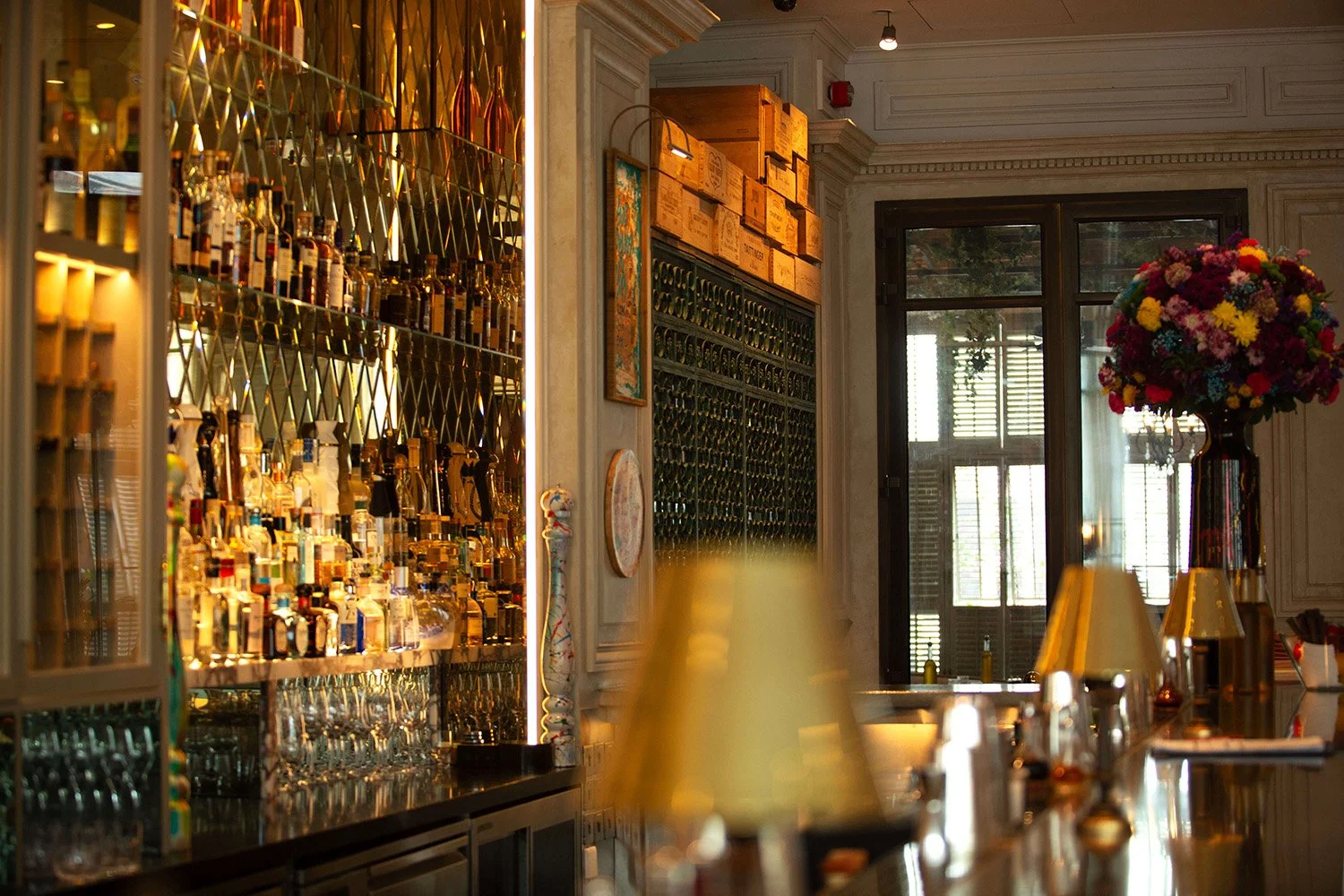 Interior of a bar or restaurant with a well-stocked alcohol shelf, bar counter with glasses and lamps, a large bouquet of colorful flowers, and windows.