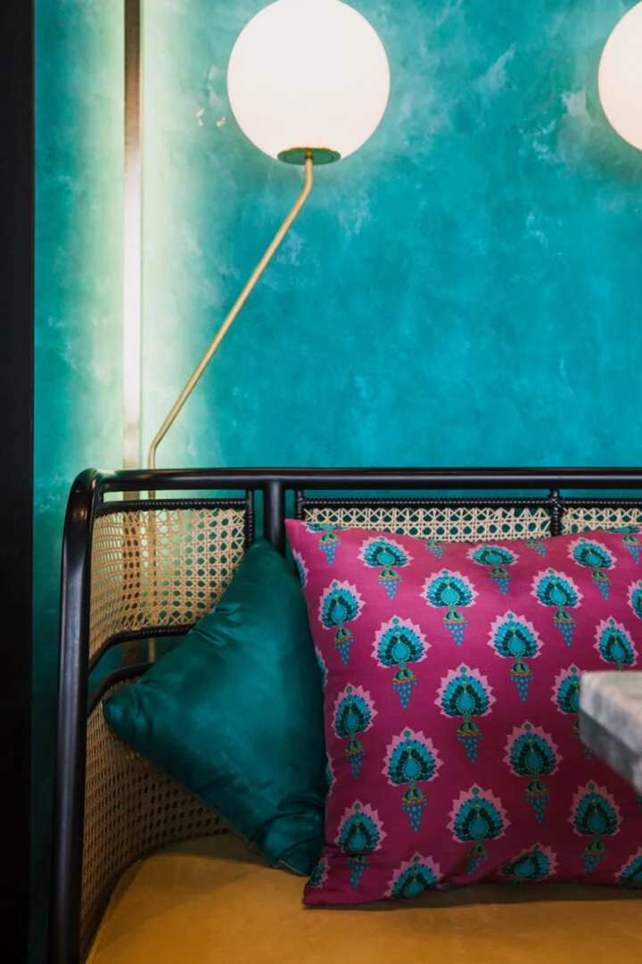 Close-up of a colorful interior at Masti restaurant with patterned pillows, a teal pillow, and a rattan headboard against a blue-green wall