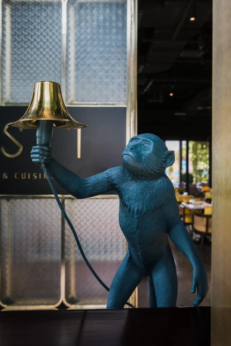 Black sculpture of a chimpanzee holding a gold-colored table lamp in a restaurant interior.