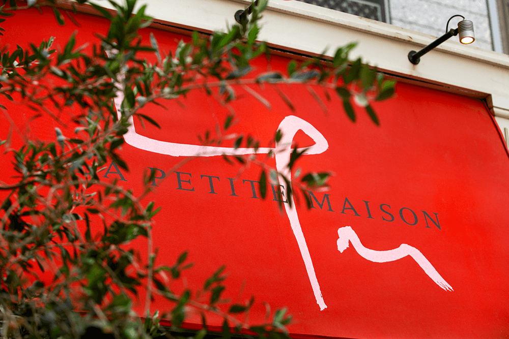 Red storefront sign with black text reading 'A PETITE MAISON' and a white abstract line drawing in the background, partially obscured by green foliage.