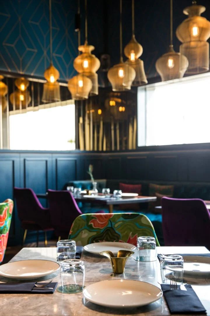 Masti restaurant table set with plates, glasses, and a gold pitcher, with colorful chairs and modern pendant lights hanging from the ceiling.