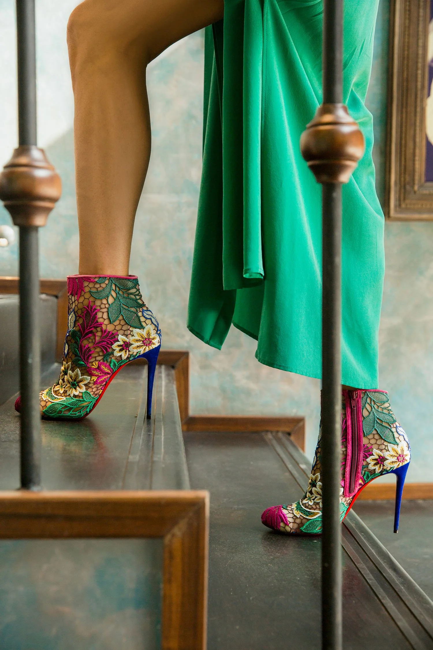 Close-up of a mannequin's legs wearing colorful embroidered high-heeled boots with floral designs, standing on a wooden staircase with a green dress flowing down.