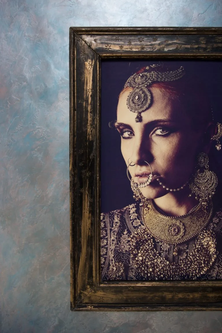 A framed portrait of a woman wearing traditional Indian jewelry and attire, with a blue textured wall in the background.