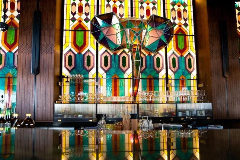 Interior of Masti restaurant with a large stained glass wall with a colorful geometric pattern behind a bar setup with glasses and bottles.