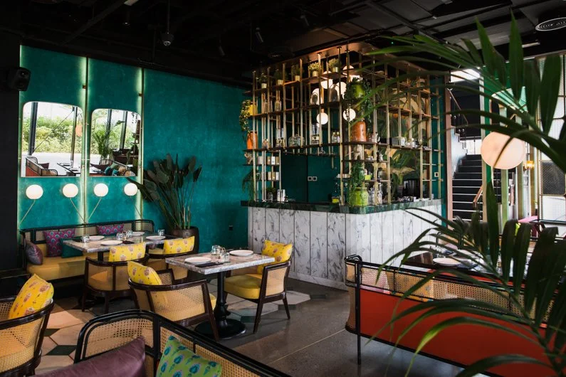 Interior of Masti restaurant in Dubai with teal walls, yellow chairs, and a marble bar with shelving and plants.