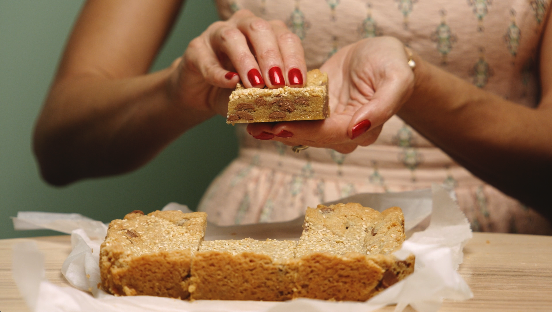 A person with red nail polish is holding a piece of protein bar over the rest of the bar, which is on a piece of parchment paper on a wooden surface.