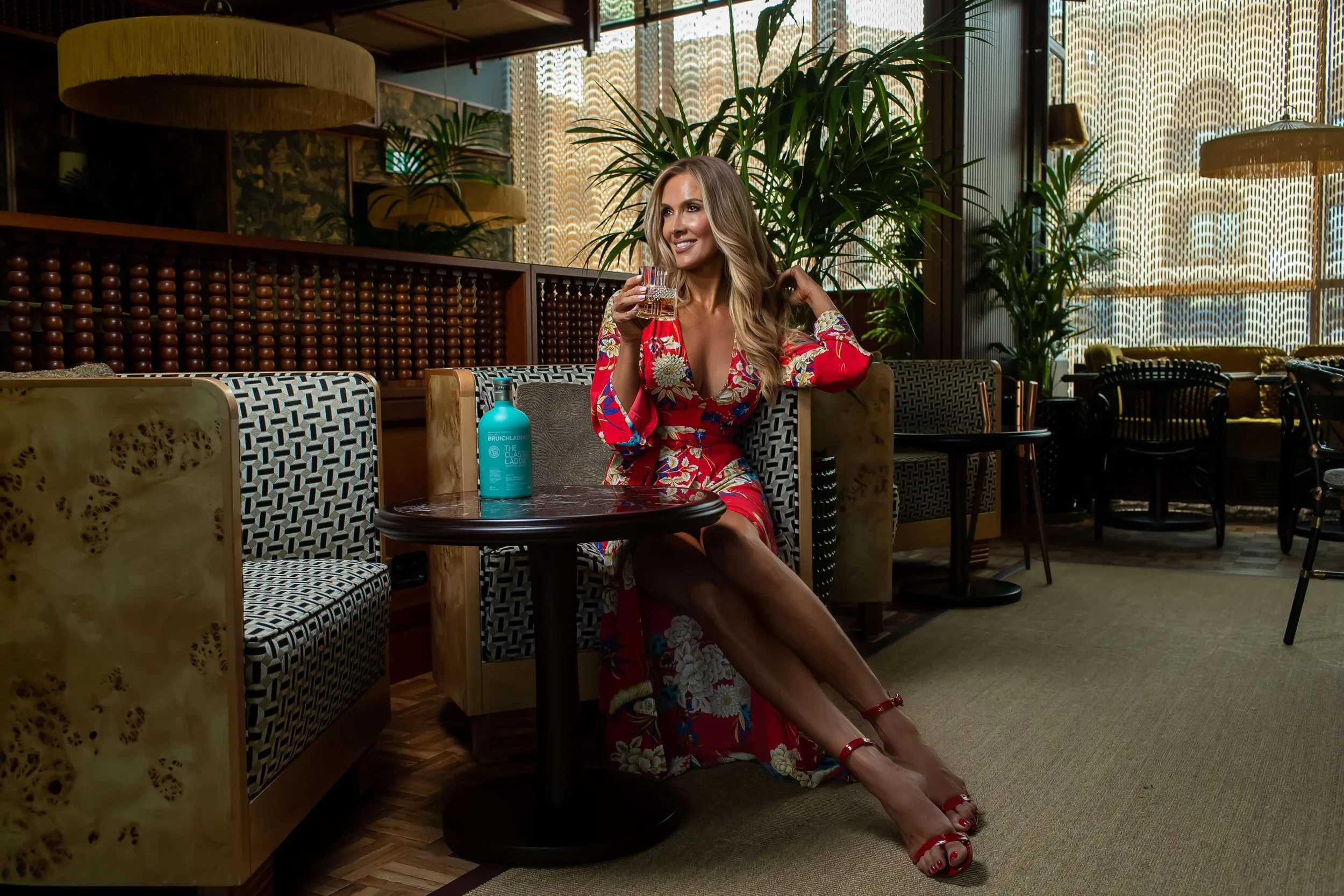 A woman in a red floral dress is sitting in a restaurant, holding a glass of drink with a smile. She has long blonde hair and wears red high heels. There is a bottle of hand sanitizer on the table in front of her, and the background features a cozy, 