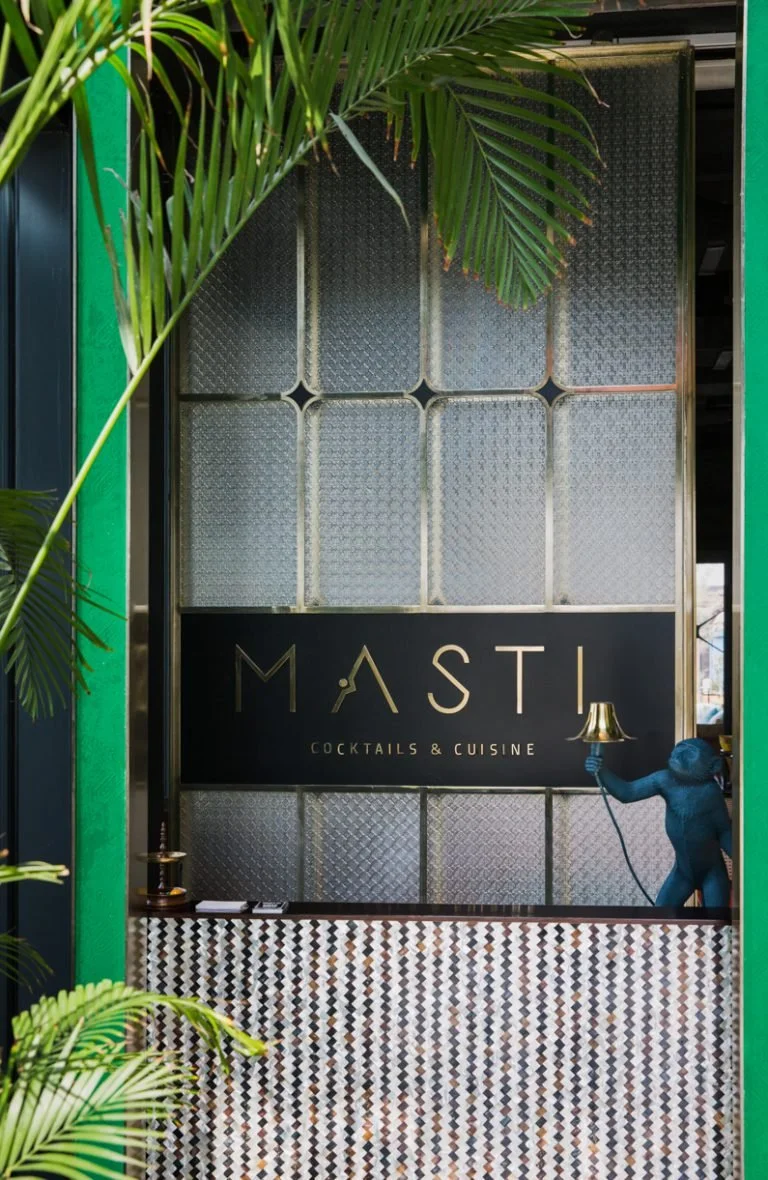 Sign for Masti Cocktails & Cuisine restaurant with a blue monkey holding a lamp inside, partially obscured by green foliage.