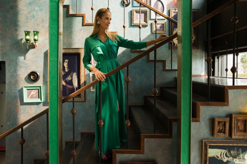 A woman in a long green dress standing on a staircase inside a decorated home with art frames on the wall.