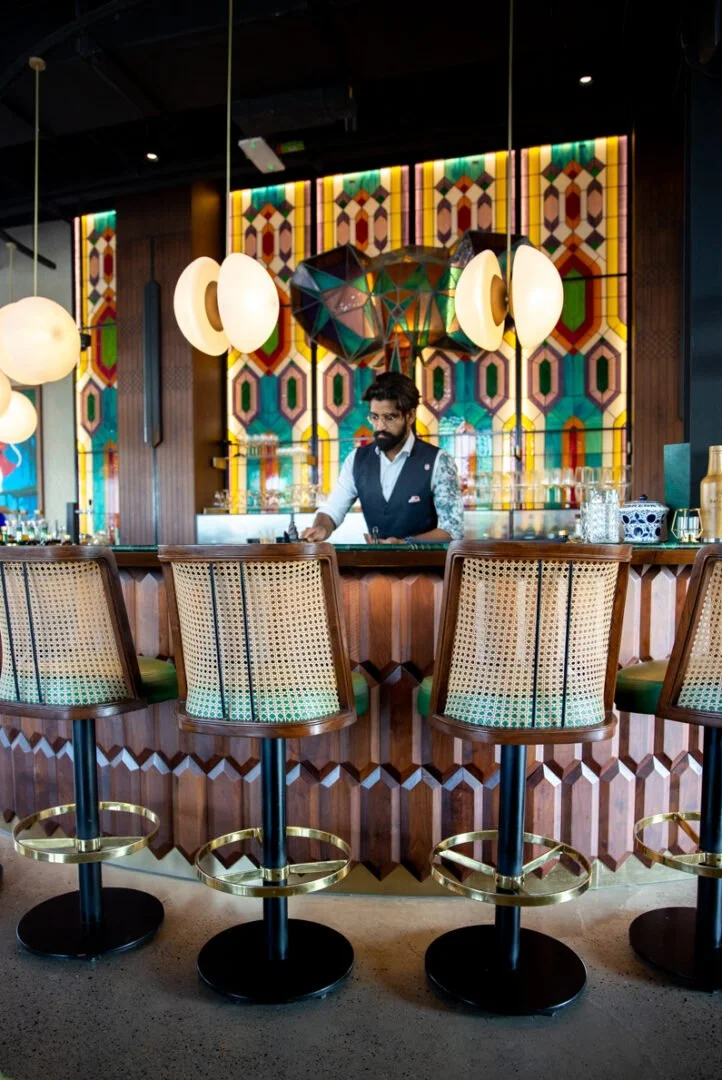 A bartender at Masti restaurant works behind a wooden bar counter, preparing a drink in a colorful, modern bar with geometric wall decor and hanging pendant lights.