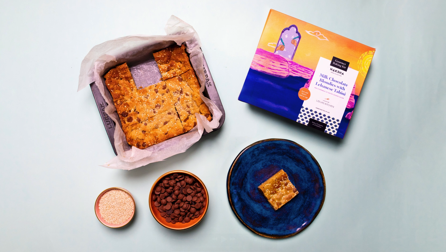 A Naksha baking kit box labeled 'Milk Chocolate Blondies with Lebanese Tahini' next to a baking tray lined with parchment paper holding sliced blondies, two bowls with chocolate chips and sprinkles, and a blue plate
