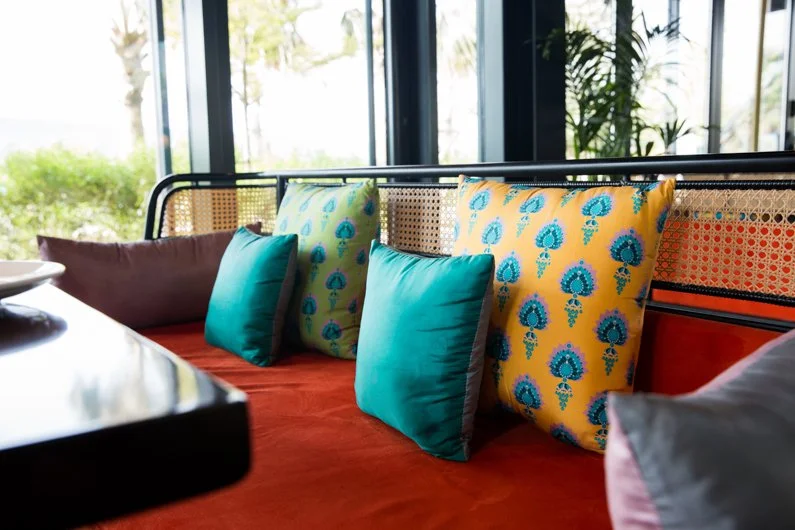 A row of colorful pillows on a red cushioned bench near large windows with a garden view.