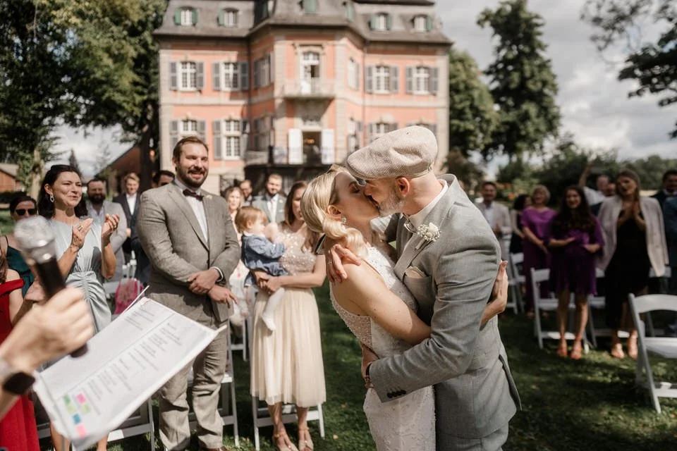 Ein Brautpaar bei einer Hochzeit, das sich küsst, umgeben von Gästen, im Freien vor einem großen, mehrstöckigen Haus.