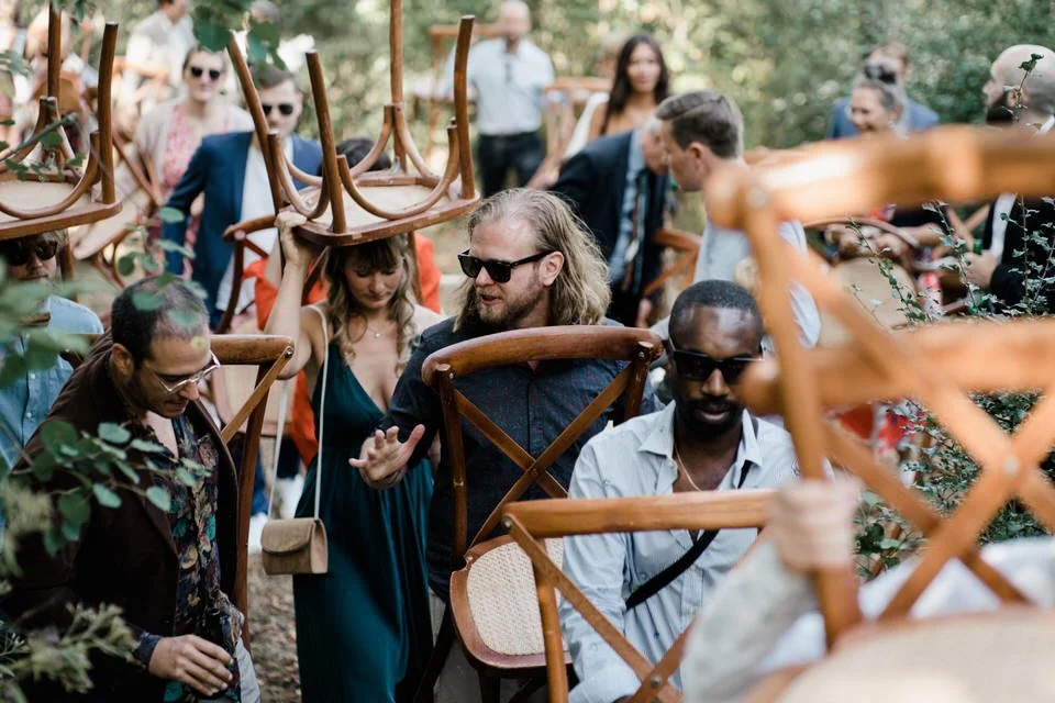 Menschen stehen im Wald, einige tragen Stühle auf den Köpfen, es scheint eine größere Veranstaltung oder Demonstration zu sein.
