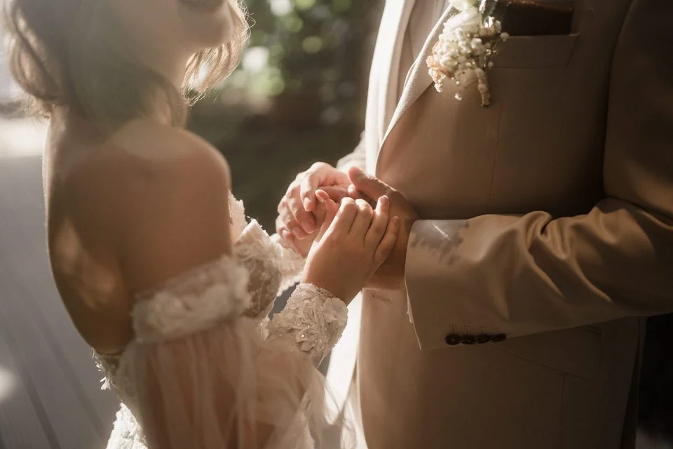 Ein Brautpaar hält sich bei einer Hochzeitshand in Hand. Die Frau trägt ein weißes, hoch geschlitztes Brautkleid mit Spitze, während der Mann einen beige Anzug trägt. Die Szene ist romantisch und emotional, wahrscheinlich während der Zeremonie.