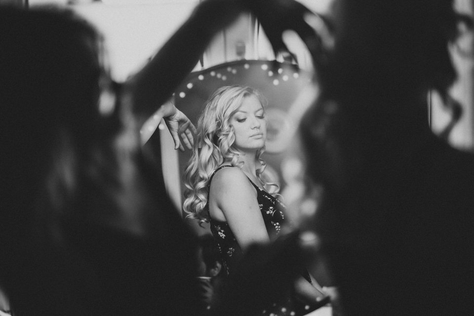 Eine junge Frau mit langen, gewellten Haaren posiert in einem Spiegel, umgeben von zwei Personen, die ihre Haare stylen.