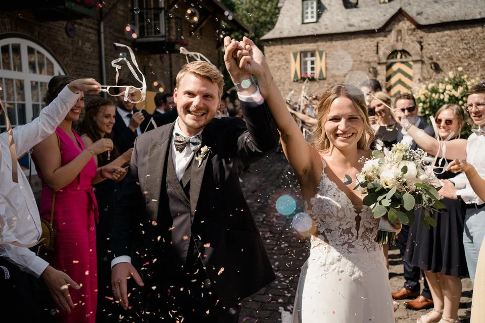 Ein glückliches Brautpaar bei ihrer Hochzeitsfeier, umgeben von Freunden, die Konfetti werfen, draußen vor einem alten Gemäüse, mit Sonnenlicht und bunter Dekoration.