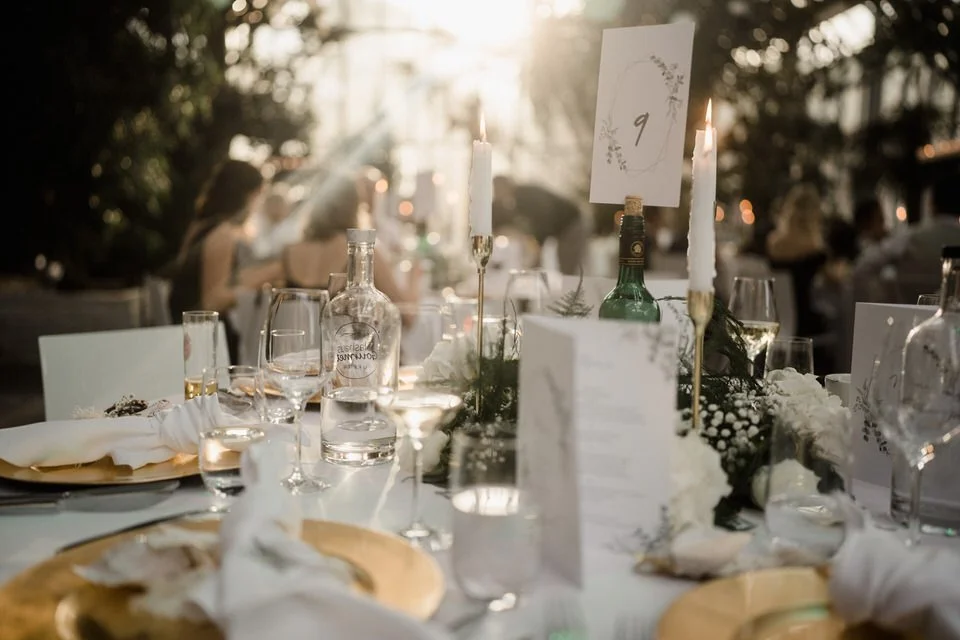Festlich gedeckter Tisch bei einer Veranstaltung im Freien, mit Kerzen, Gläsern, Weingläsern, einer Flasche und Menükarten, bei Sonnenuntergang.