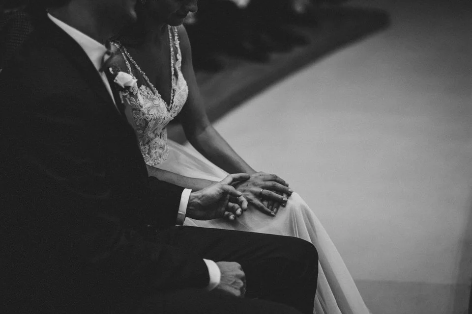 Braut und Bräutigam sitzen nebeneinander bei einer Hochzeit, die Hände sind ineinander verschränkt, in einem schwarzen und weißen Foto