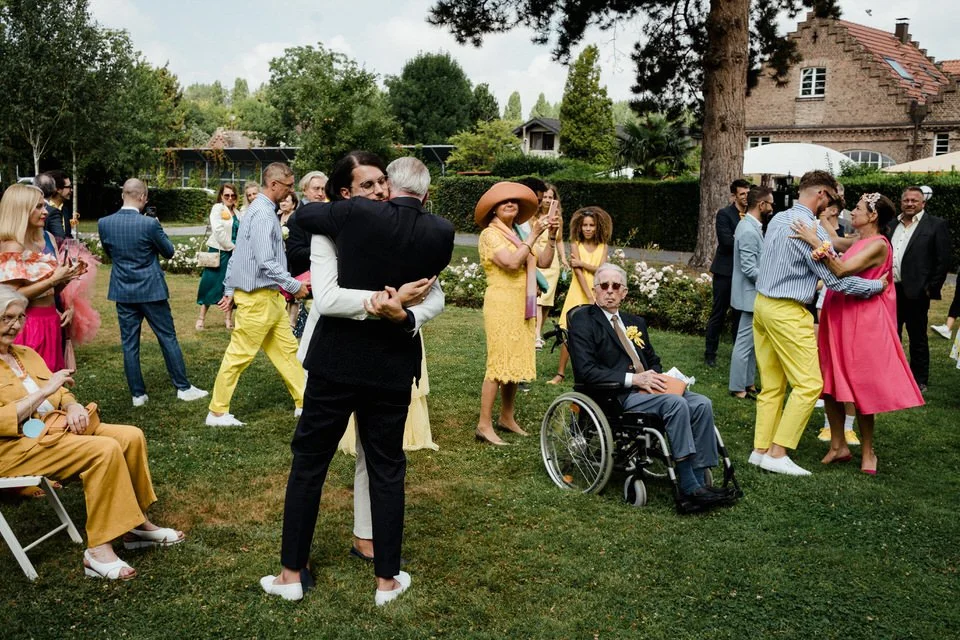 Eine Gruppe von Menschen bei einer Hochzeit im Garten, einige tanzen, andere fotografieren oder sitzen, inklusive einem älteren Mann im Rollstuhl, und alle sind gekleidet in festliche Kleidung.