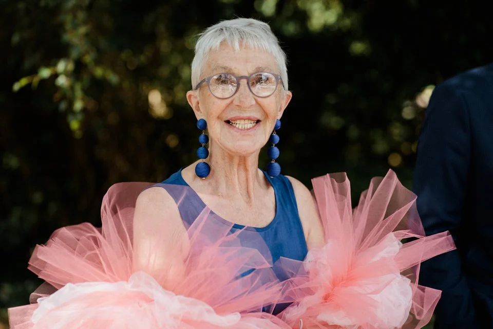 Lächelnde ältere Frau mit kurzen grauen Haaren, großen runden Brillen und blauer Kleidung, umgeben von rosa Tüll bei einer Feier im Freien.