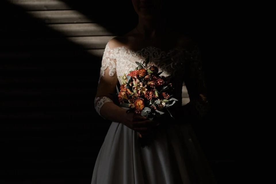 Eine Braut in einem weißen Kleid hält einen Blumenstrauß, im Schatten, nur teilweise sichtbar.