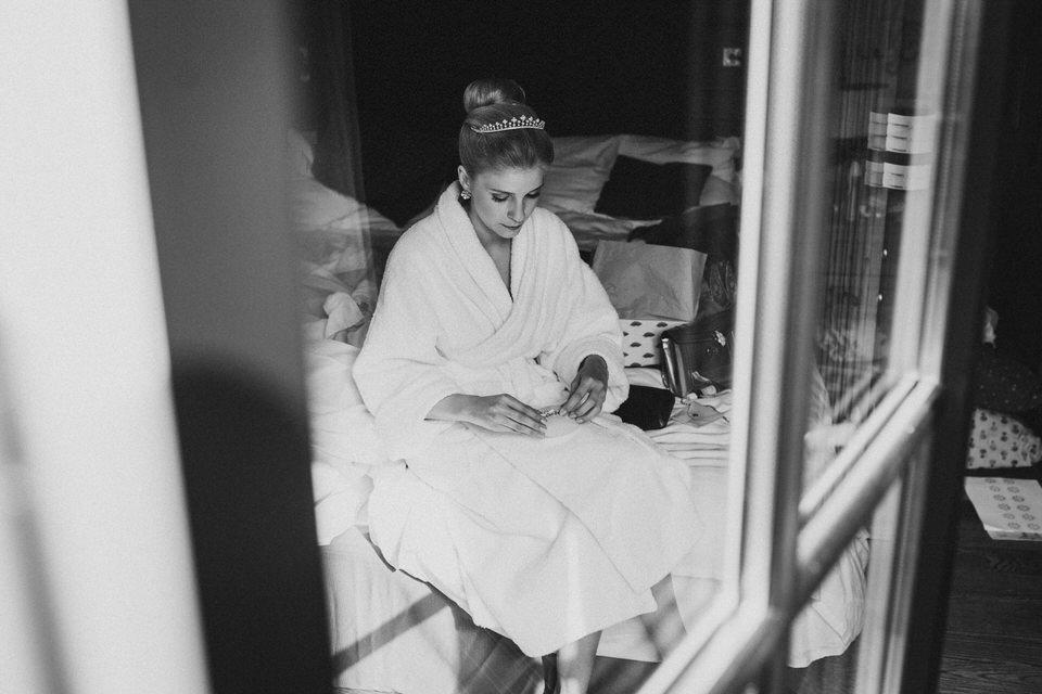 Eine junge Frau in einem Bademantel mit einer Krone auf dem Kopf sitzt auf einem Bett, betrachtet ihr Smartphone, im Hintergrund sind Kissen und Bettwäsche zu sehen.