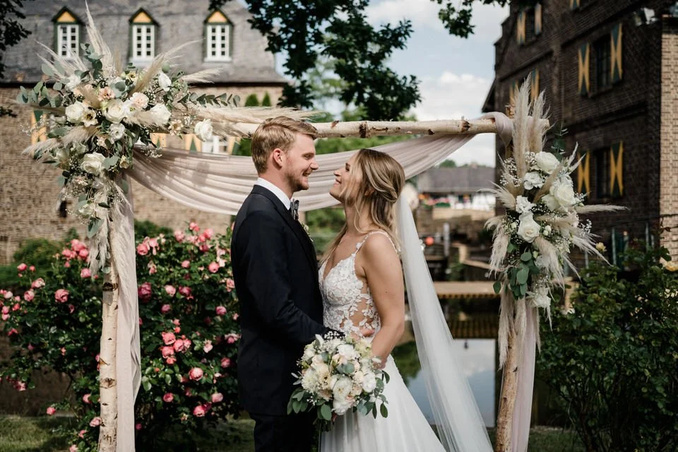 Ein Brautpaar steht sich bei ihrer Hochzeit gegenüber unter einem dekorierten Holzbogen mit weißen Blumen und Stoffen, umgeben von blühenden Rosen im Garten, vor historischen Gebäuden.
