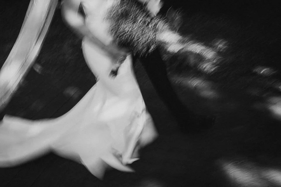 Ein Hund, der einen schwarzen Gegenstand im Maul hält, steht auf einem Holzboden. Der Hund trägt ein weißes Kleidungsstück.