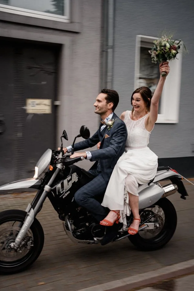 Ein Brautpaar fährt auf einem Motorrad, die Braut hält einen Blumenstrauß hoch, beide scheinen glücklich.