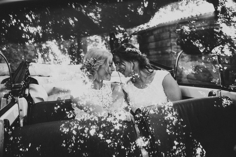 Zwei Frauen in Brautkleidern, die sich mit ihren Stirnflächen berühren, sitzend im offenenAuto, draußen mit Bäumen im Hintergrund, in Schwarz-Weiß aufgenommen.