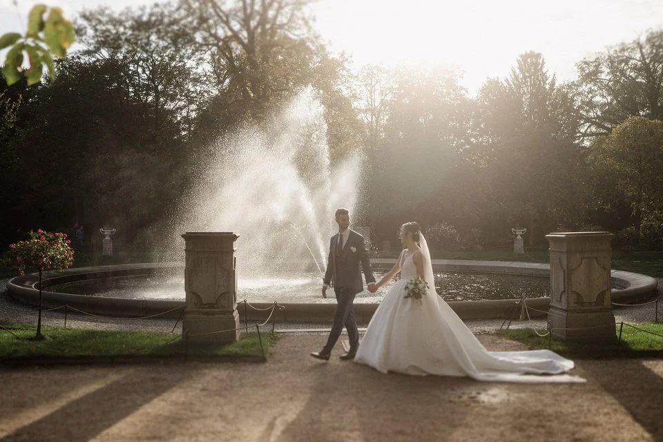 Ein Brautpaar bei Sonnenuntergang vor einem Brunnen im Park, die Braut in einem weißen Kleid und der Bräutigam in einem dunklen Anzug, Hand in Hand.