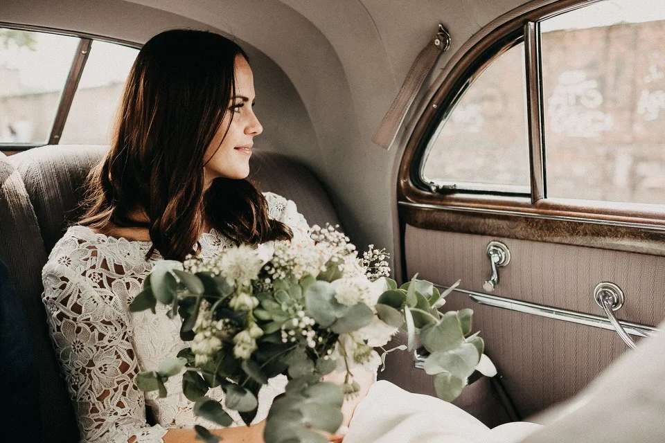 Eine Frau in einem weißen Spitzenkleid sitzt im Fond eines Autos, hält einen Blumenstrauß und schaut nach draußen.