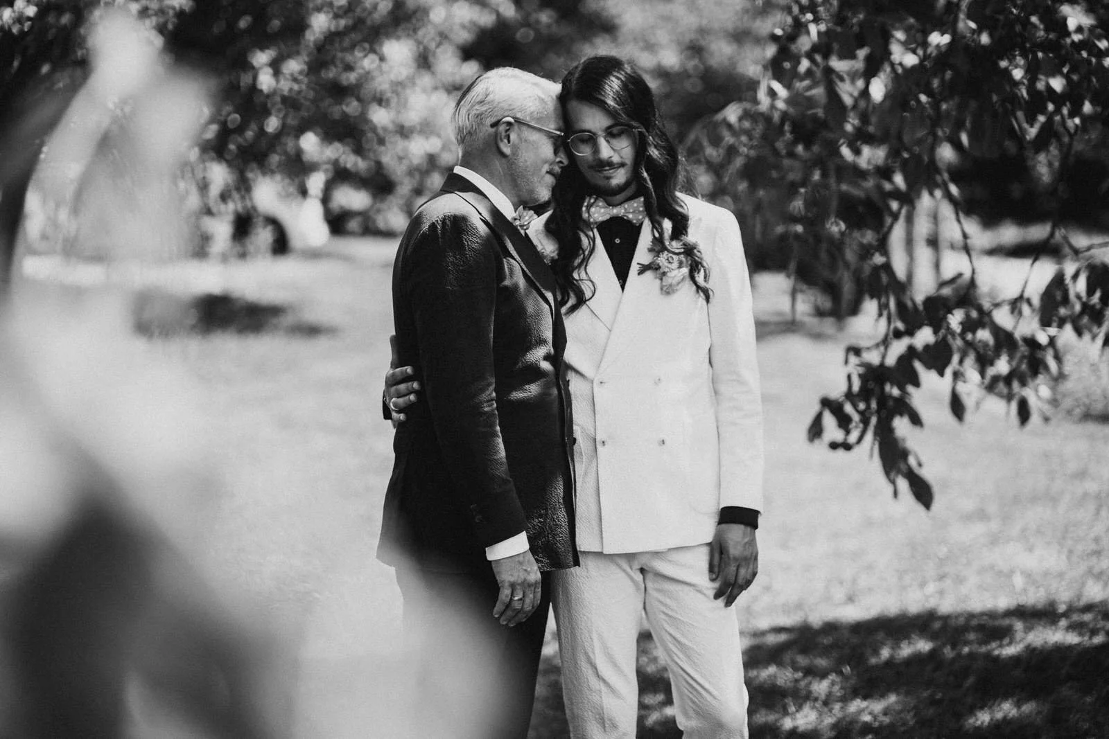 Zwei Männer, einer älter, der andere jünger, stehen eng beieinander im Grünen. Der Jüngere trägt einen weißen Anzug mit Blumen und einen schwarzen Hemd, der Ältere trägt einen schwarzen Anzug. Es ist ein Schwarz-Weiß-Foto, vermutlich bei einer formel