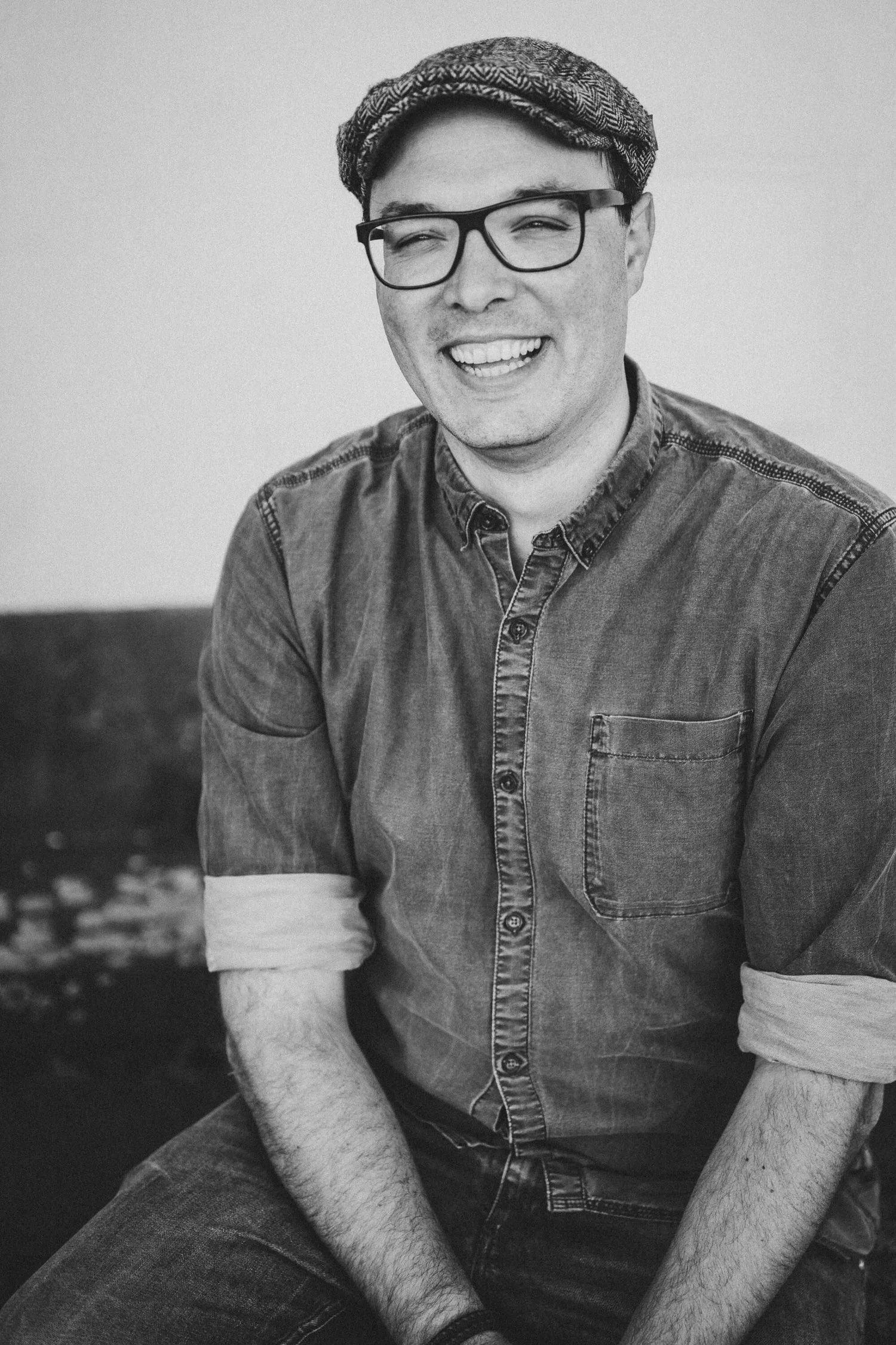 Moritz. Ein lachender junger, gut gelaunter Mann mit Brille, Mütze und Jeanshemd.