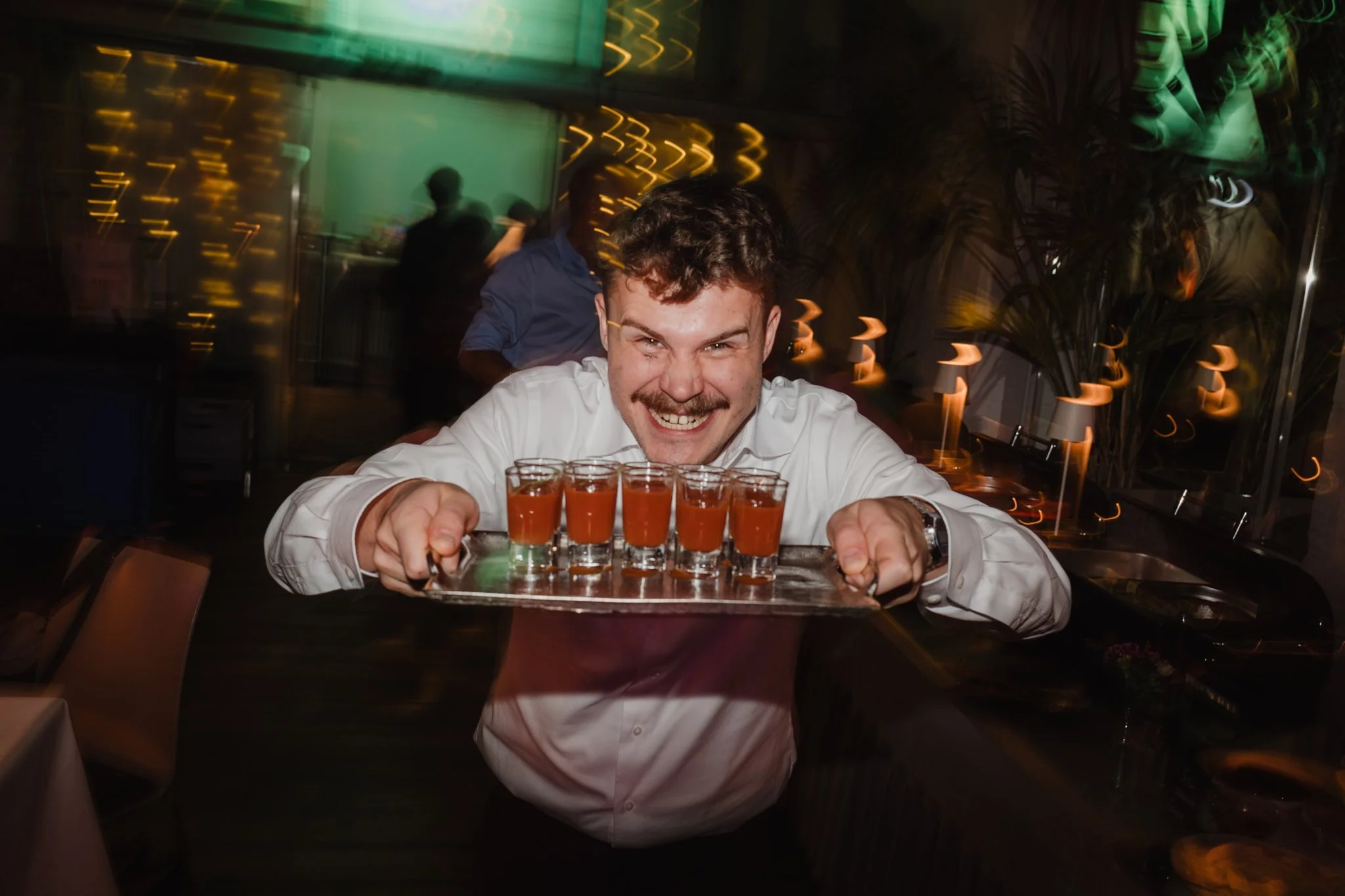 Ein fröhlicher Barkeeper hält ein Tablett mit mehreren kleinen Gläsern mit orangefarbigen Getränken in einem Restaurant oder Bar bei Nacht.
