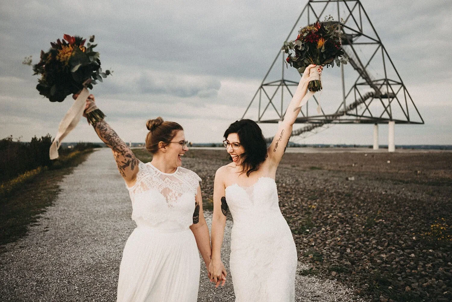 Zwei Frauen in weißen Brautkleidern feiern auf einer ländlichen Straße, halten sich an den Händen und heben ihre Blumensträuße in die Luft, während im Hintergrund eine Windkraftanlage steht.