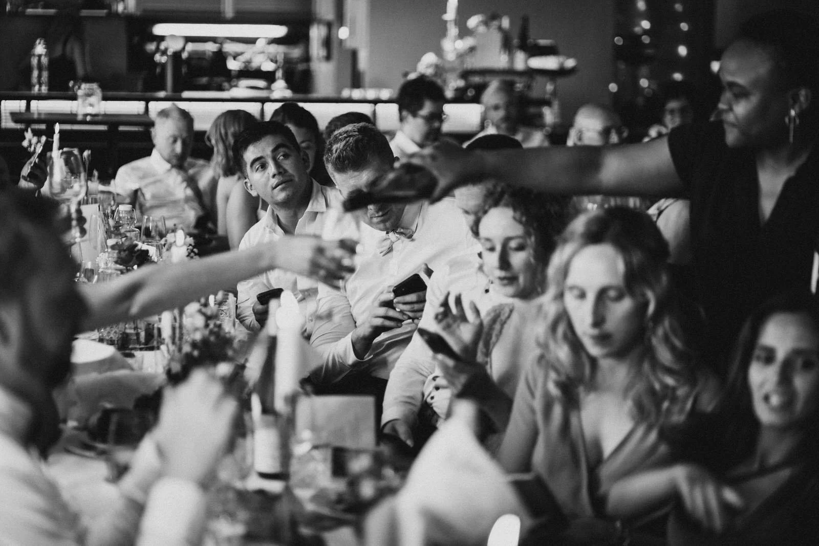 Menschen sitzen an einem Tisch in einem Restaurant oder einer Feier, einige schauen auf ihre Handys, während eine Kellnerin Getränke serviert.