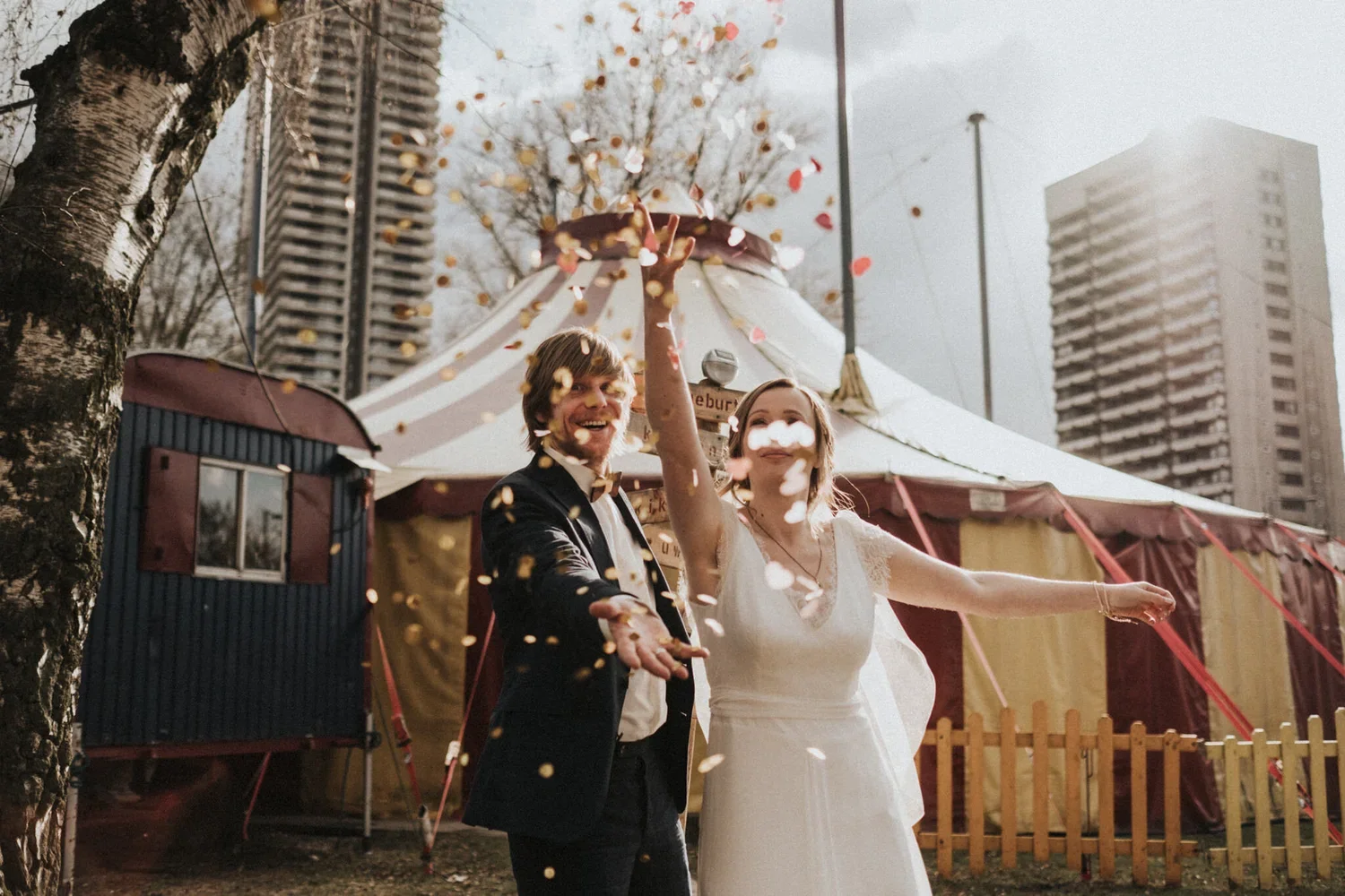 Ein glückliches Brautpaar wirft Konfetti vor einem Zirkuszelt bei einer Hochzeit im Freien, umgeben von Hochhäusern.