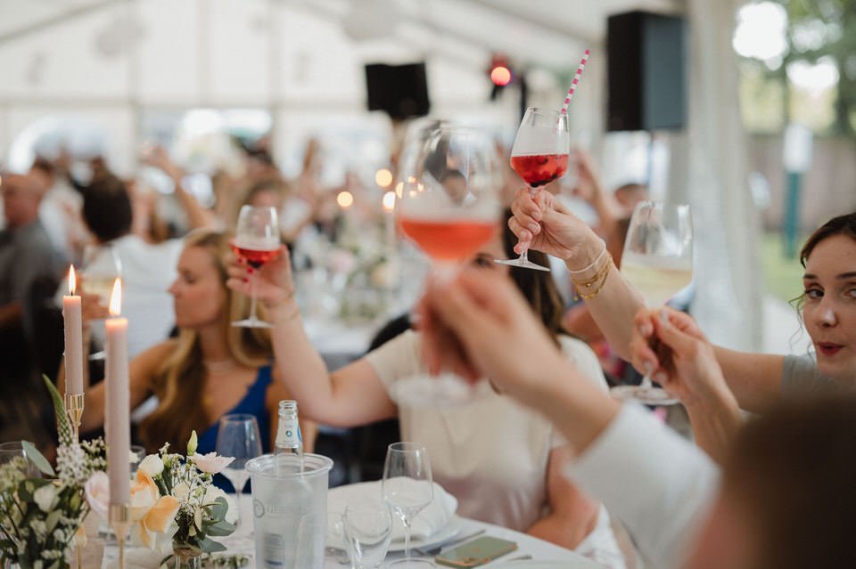 Menschen feiern mit Wein in einem festlichen Raum, viele heben ihre Gläser zum Anstoßen, es gibt Blumendekoration und Kerzen auf dem Tisch.
