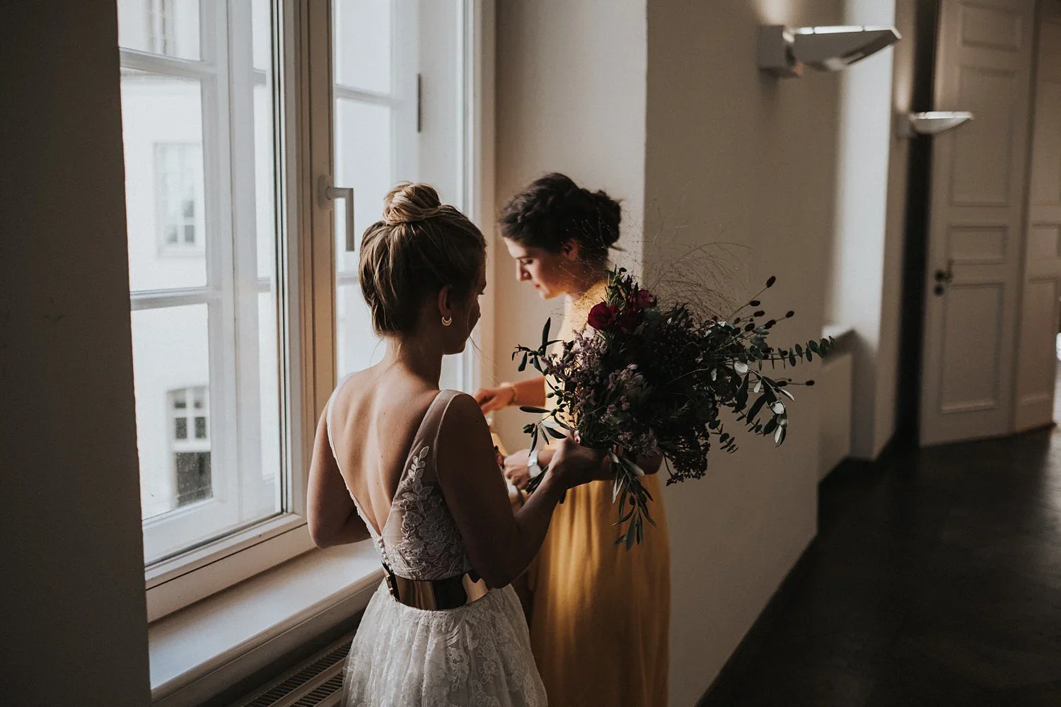 Zwei Frauen, eine im Brautkleid mit einem Brautstrauß, stehen an einem Fenster und schauen sich liebevoll an.