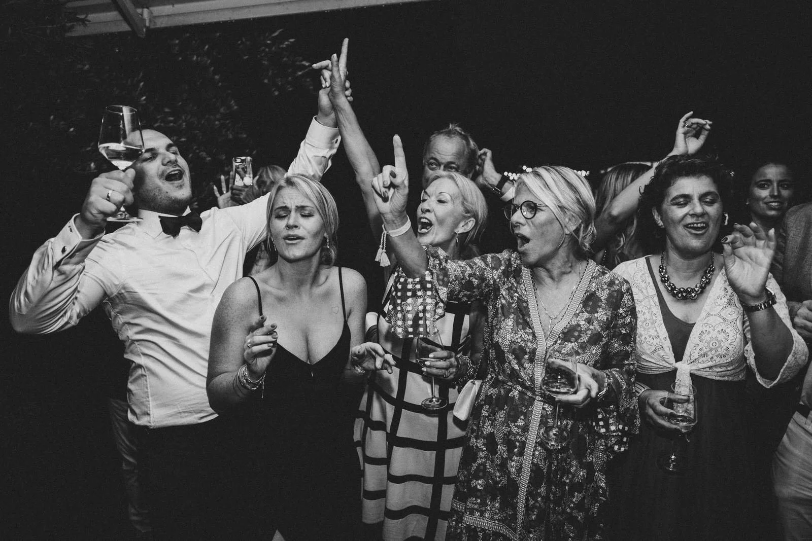 Menschen feiern und tanzen auf einer Party, einige halten Gläser mit Wein, in einer festlichen Atmosphäre bei Nacht.