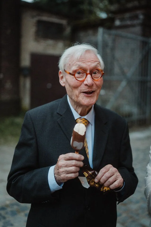 Ein älterer Mann in Anzug mit Brille und Krawatte hält ein Eis in der Hand und lächelt.
