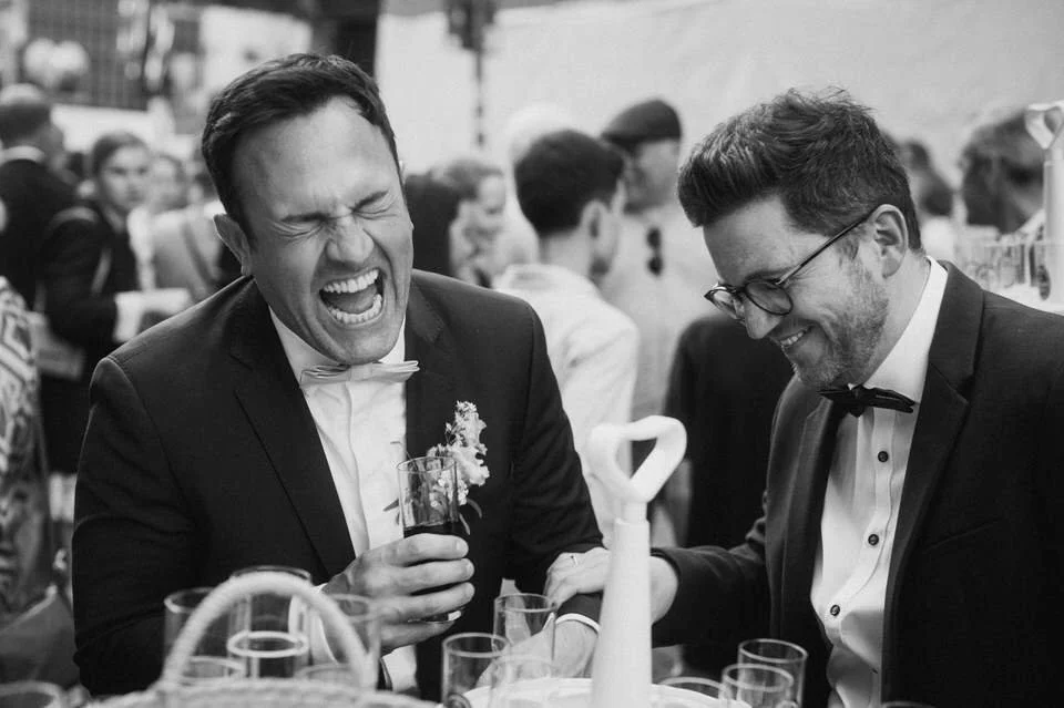 Zwei Männer in formeller Kleidung, die bei einem festlichen Anlass lachen und trinken. Im Hintergrund sind weitere Personen sichtbar.