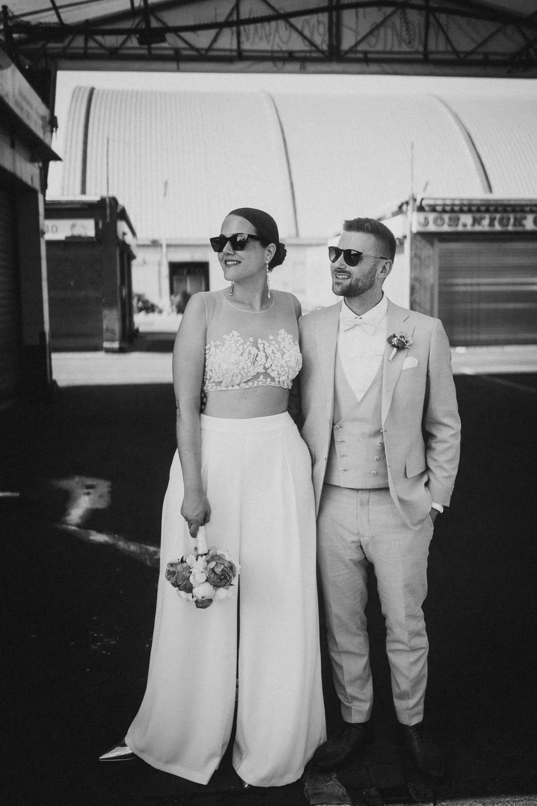 Ein Paar in formeller Kleidung steht zusammen, lächelt und trägt Sonnenbrillen. Es handelt sich um eine Hochzeit im Freien, im Hintergrund sind Buden und ein Zeltdach sichtbar.