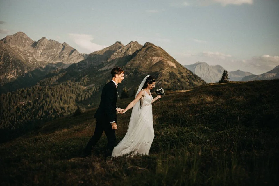 Ein Brautpaar geht Hand in Hand durch eine grüne, hügelige Landschaft mit Bergen im Hintergrund, die bei Sonnenuntergang beleuchtet wird.