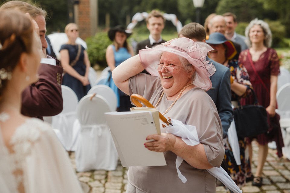 Lächelnde ältere Frau bei einer Hochzeit, trägt einen großen Hut und hält ein Geschenk, inmitten von Gästen im Freien.